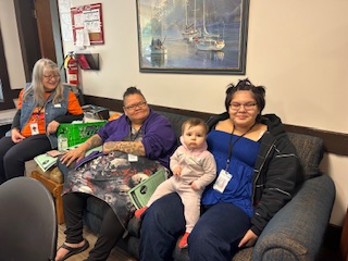 Four women and a baby sit together on a couch during a retreat gathering. One woman holds the baby on her lap, and all appear relaxed and smiling. 