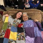 Two women sit closely together on a couch, smiling warmly as they wrap themselves in colourful handmade quilts. Other retreat participants can be seen in the background, engaged in conversation and activities.